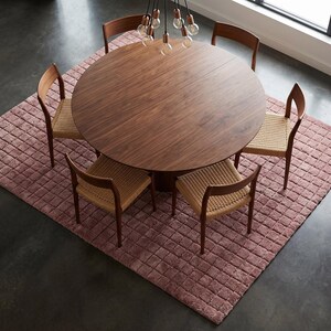 May include: A round dark wood dining table with a central pedestal base, surrounded by five wooden chairs featuring woven tan seats. The furniture set is arranged on a textured, square, dusty rose rug with a grid pattern on a dark floor.