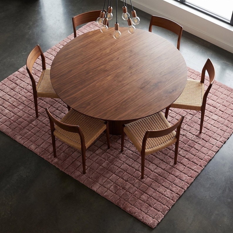 May include: A round dark wood dining table with a central pedestal base, surrounded by five wooden chairs featuring woven tan seats. The furniture set is arranged on a textured, square, dusty rose rug with a grid pattern on a dark floor.