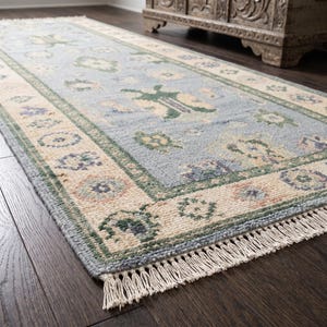 May include: A rectangular area rug featuring a light blue central field with green, cream, and purple floral motifs. The rug has a wide cream-colored border with intricate patterns and is finished with short white fringe along the short edges.