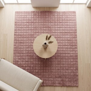 May include: A large, rectangular, dusty rose area rug featuring a grid of raised, square-shaped tufted sections. The rug is positioned on a light-colored wooden floor, with a round wooden coffee table and a small vase with dried grass on top.