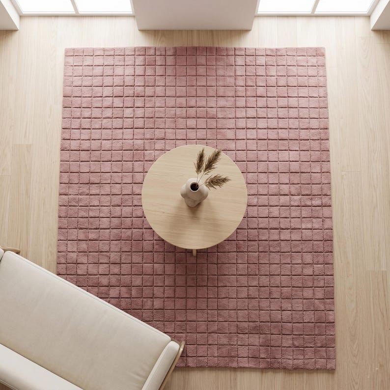 May include: A large, rectangular, dusty rose area rug featuring a grid of raised, square-shaped tufted sections. The rug is positioned on a light-colored wooden floor, with a round wooden coffee table and a small vase with dried grass on top.