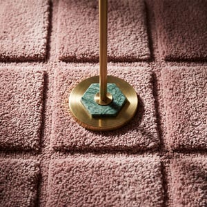 May include: A dusty rose rug with a grid of raised square tufted sections. Centered on the rug is a circular gold metal base with a thin vertical gold rod extending upward, featuring a hexagonal green marble inlay at the connection point.