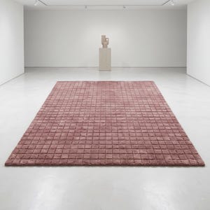May include: A rectangular area rug in a dusty rose color featuring a uniform grid pattern of small, raised square tufts across the entire surface. The rug is laid flat on a smooth, light-colored floor in a large, open, white-walled gallery space.
