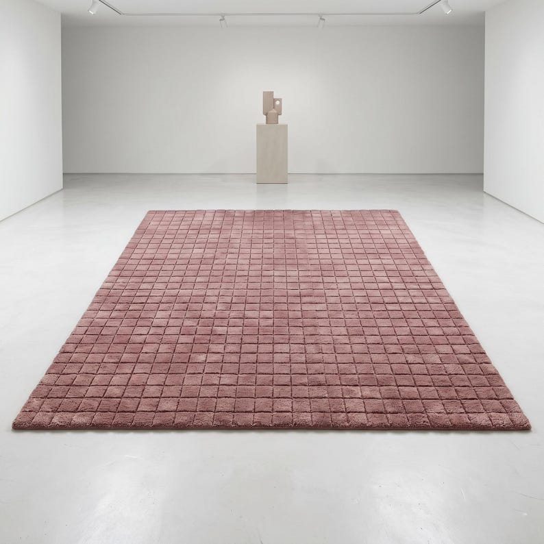 May include: A rectangular area rug in a dusty rose color featuring a uniform grid pattern of small, raised square tufts across the entire surface. The rug is laid flat on a smooth, light-colored floor in a large, open, white-walled gallery space.