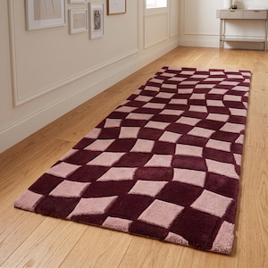 Puede incluir: Una alfombra rectangular con un patrón de tablero de ajedrez distorsionado y ondulado en tonos alternos de burdeos intenso y rosa empolvado. La alfombra tiene una textura gruesa y tupida, colocada sobre un suelo de madera clara.
