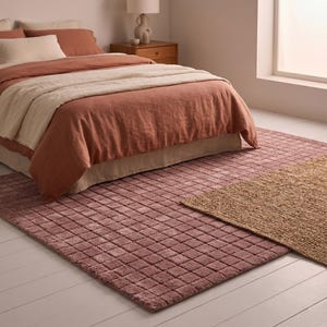 May include: A rectangular area rug featuring a raised grid pattern in a dusty rose color, layered partially beneath a second rectangular rug made of natural tan woven fibers. The rugs are positioned on a white floor in front of a bed.
