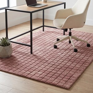 May include: A rectangular area rug featuring a textured, grid-like pattern of raised squares in a dusty rose color. The rug is positioned beneath a desk with a black metal frame and a light wood top, and a cream-colored swivel office chair with wheels.