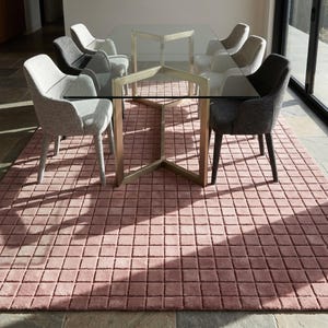 May include: A rectangular area rug with a raised grid pattern in a dusty rose color. The rug is positioned under a glass-topped dining table with a gold-toned metal base and six upholstered armchairs in shades of light gray and dark charcoal.