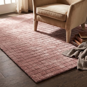 May include: A rectangular area rug in a dusty rose pink hue featuring a raised, grid-like texture of small, uniform squares. The rug is positioned on a dark wood floor, partially underneath a beige upholstered armchair and a stack of three books.