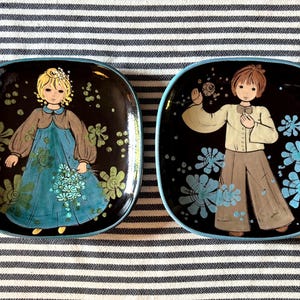 May include: Two square ceramic plates with rounded corners and a black background. The left plate features a blonde figure in a blue dress holding flowers. The right plate shows a figure in a light shirt and brown trousers with blue floral accents.