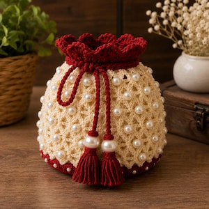 Peut inclure: Une pochette à cordon en crochet de couleur crème avec un motif en losanges répété orné de perles blanches. Le sac présente un bord festonné rouge foncé et un cordon de serrage rouge assorti terminé par deux pompons rouges et deux perles.