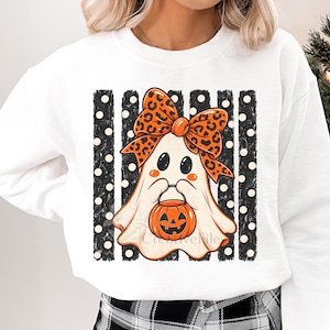 May include: A white crewneck sweatshirt featuring a graphic of a ghost wearing a large orange and black leopard print bow. The ghost holds a small orange jack-o'-lantern bucket, set against a background of vertical black stripes with white polka dots.