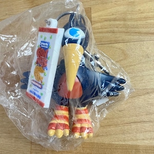 Pode incluir: Um porta-chaves de pássaro azul-escuro com um bico amarelo grande, um olho azul e pés às riscas cor de laranja e amarelas. Preso ao anel metálico está um pendente retangular branco com o gráfico de um saco de Corn Nuts.