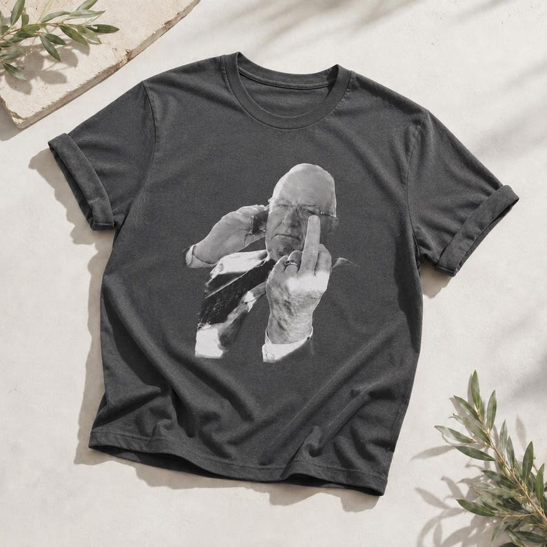 May include: A charcoal gray short-sleeve t-shirt featuring a black and white graphic print of a person wearing glasses, holding a phone to their ear, and gesturing with their middle finger raised. The shirt has a crew neckline and rolled sleeves.