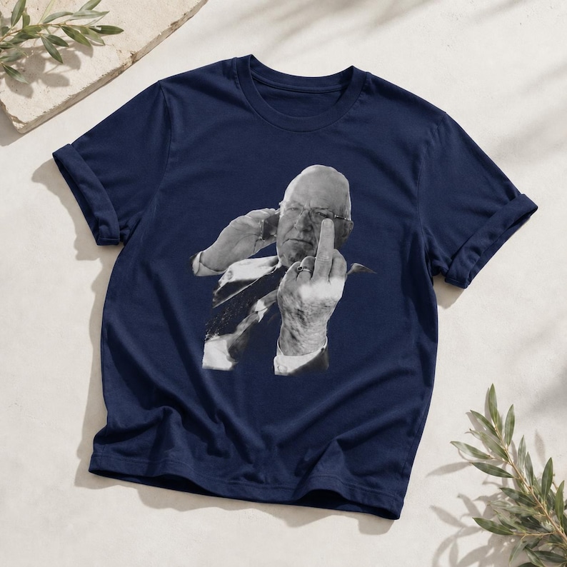 May include: A navy blue short-sleeve t-shirt featuring a black and white graphic print of a person wearing glasses, holding a phone to their ear, and gesturing with their middle finger raised toward the viewer in a prominent display.