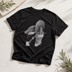 May include: A black short-sleeved t-shirt featuring a grayscale photographic print of an older person wearing glasses and a suit, holding up their middle finger. The graphic is centered on the chest of the shirt, which has a crew neckline.