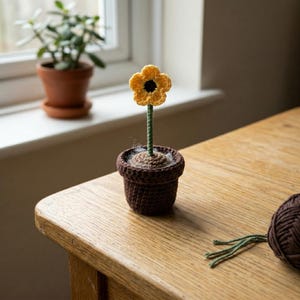 May include: A small crocheted flower with yellow petals and a black center, attached to a green stem. The flower is planted in a dark brown crocheted pot with a tan top surface. A ball of dark brown yarn with green yarn ends sits nearby.