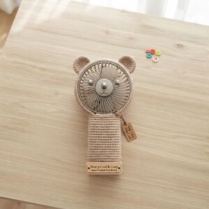 Puede incluir: Un ventilador portátil de mano con una funda de ganchillo beige en forma de oso con orejas redondeadas. El ventilador tiene una rejilla metálica y una etiqueta de madera que dice Stay Cool. Una base de madera rectangular lleva la etiqueta Bear-y Cool & Cozy.