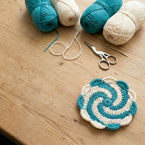 Op de afbeelding: Een ronde, gehaakte onderzetter met een spiraalpatroon in blauwgroen en crèmekleurig garen. Daarnaast liggen twee bollen blauwgroen garen, twee bollen crèmekleurig garen, een zilverkleurige metalen schaar en een enkele metalen naainaald.
