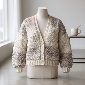 May include: A long-sleeve V-neck cardigan featuring a textured bobble stitch pattern in a colour-blocked design of cream, light grey, and dark grey. The garment includes a ribbed hem and cuffs, and is fastened with three round, light-coloured buttons.
