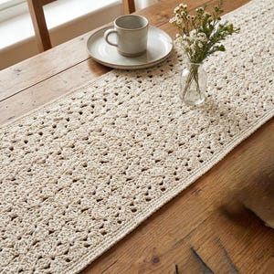 Puede incluir: Un camino de mesa rectangular de ganchillo en color crema con un patrón de motivos florales repetitivos. El camino está extendido en plano, mostrando el intrincado trabajo de hilo y la textura del diseño textil hecho a mano en toda su longitud.