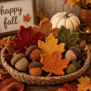 May include: A woven basket filled with crocheted autumn decor, including maple leaves in red, orange, yellow, and green, alongside several crocheted acorns with brown caps and tan or orange bodies, arranged with a sign reading happy fall.