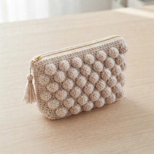 May include: A beige crochet pouch featuring a textured bobble stitch pattern across the exterior. The rectangular bag includes a gold-toned metal zip closure along the top edge and a matching beige yarn tassel attached to the zip pull.