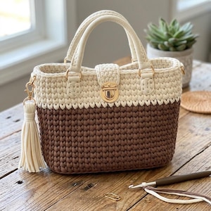 May include: A two-toned crochet handbag featuring a cream-coloured upper section and a dark brown lower section. The bag includes two cream handles, a gold-toned metal turn-lock closure, and a cream-coloured tassel attached to a gold-toned side clip.