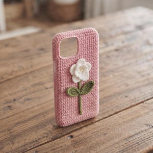 Può includere: Una custodia per telefono all'uncinetto di colore rosa con un fiore bianco, uno stelo verde e due foglie al centro. La custodia presenta un'apertura rettangolare nella parte superiore per l'obiettivo della fotocamera e una trama fitta.