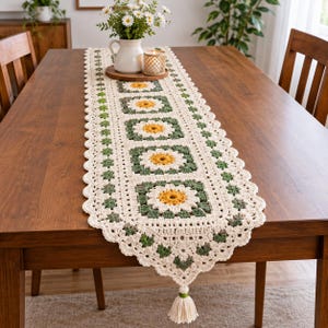 May include: A rectangular crochet table runner featuring a repeating pattern of square motifs with yellow and white daisy designs surrounded by dark green borders. The runner is finished with an off-white scalloped edge and a decorative tassel.