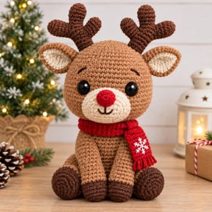 Puede incluir: Un reno de peluche tejido a ganchillo con pelaje marrón claro, astas y pezuñas de color marrón oscuro. Presenta grandes ojos negros, una nariz roja brillante y una bufanda roja tejida con un detalle de copo de nieve blanco bordado.