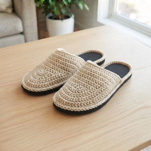 May include: A pair of beige crochet slippers featuring a textured, circular stitch pattern on the upper part and a solid black sole. The slippers have an open-back design with a rounded toe shape, showcasing a consistent yarn weave throughout.