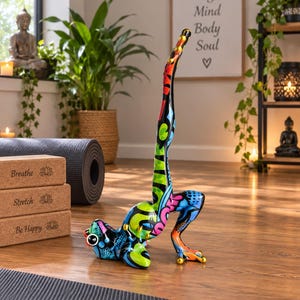 May include: A colourful frog figurine in a handstand pose, featuring vibrant graffiti-style patterns in blue, green, red, and orange. The frog has large eyes and gold-coloured accents on its toes, standing on a wooden floor near three stacked cork yoga blocks.