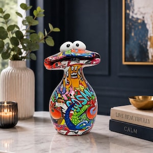 May include: A decorative ceramic figurine shaped like a stylised creature with a wide, flat head and two large, round white eyes. The body is covered in a vibrant, multi-coloured graffiti-style pattern featuring various abstract shapes and text.