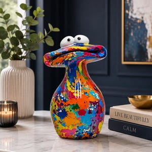 May include: A decorative figurine shaped like a stylised creature with a wide, flat head and two large, white, round eyes. The body features a vibrant, multi-coloured paint splatter pattern in shades of blue, orange, yellow, red, and green.