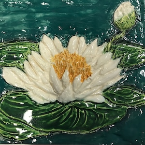 Puede incluir: Azulejo de cerámica con un nenúfar blanco con centro amarillo, rodeado de hojas de nenúfar verdes y un capullo. El fondo es un efecto de agua brillante de color verde azulado. El azulejo tiene un aspecto texturizado y tridimensional.