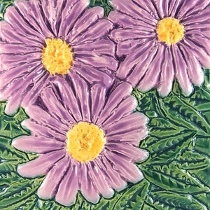 May include: Close-up of three purple and yellow flowers with green leaves. The flowers have a 3D effect and are painted on a ceramic tile.