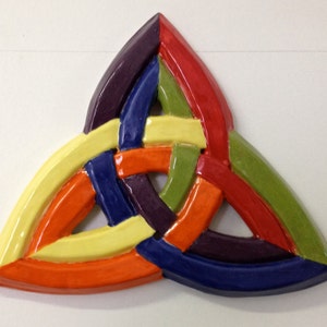 May include: A colorful ceramic wall hanging in the shape of a Celtic knot. The knot is made of five interwoven loops, each painted a different color: red, orange, yellow, green, and purple.