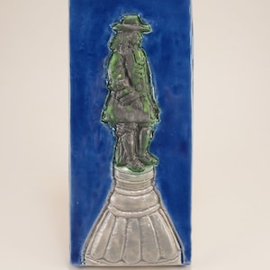 May include: A ceramic tile with a blue background and a raised image of a person in a green coat and hat standing on a white pedestal.