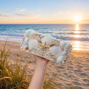 May include: A rectangular clutch bag covered in an assortment of white seashells, a central tan starfish, and scattered iridescent pink and blue pearls. The surface is densely decorated with various shell shapes and sizes, creating a textured exterior.