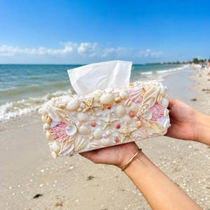 May include: A rectangular tissue box cover decorated with an assortment of white and pink seashells, starfish, and small white pearls. The exterior is completely covered in various shell textures and shapes, with a white tissue protruding from the top.