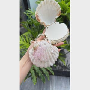 Könnte beinhalten: Ein muschelförmiges Notizbuch mit einer weißen Außenseite, die mit rosa Glitzer, kleinen weißen Perlen und klaren Kristallen verziert ist. Das Notizbuch hat ein Klappdesign mit einem Perlenhenkel und enthält mehrere leere weiße Seiten.