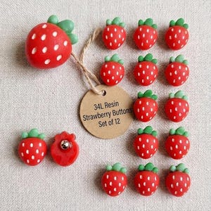 May include: A set of 12 red strawberry-shaped buttons with green leaves and white dots. Each button features a shank back. A round tan tag is attached with twine, printed with the text 34L Resin Strawberry Buttons Set of 12. The buttons are arranged in a grid pattern on a textured fabric surface.