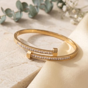 Puede incluir: Un brazalete de metal en tono dorado con un diseño inspirado en un clavo y dos extremos planos y cuadrados. La superficie exterior del brazalete está incrustada con una fila continua de pequeñas piedras transparentes de corte rectangular.