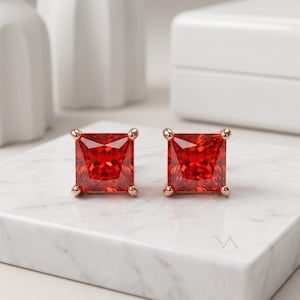 May include: A pair of square-cut, vibrant red gemstone stud earrings set in rose gold metal. Each earring features a faceted princess-cut stone secured by four prongs, displaying a deep crimson hue and a reflective, polished surface finish.
