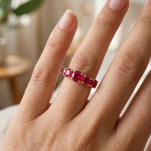 Può includere: Un anello in oro con cinque pietre preziose rotonde di un rosso vibrante disposte in fila. La pietra più grande si trova al centro, affiancata da due pietre più piccole su ciascun lato, fissate da piccole griffe dorate sulla fascia.