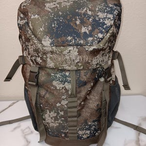 May include: A camouflage backpack featuring a brown, green, and grey pattern with the text RED HEAD 1856 embroidered on the front flap. The bag includes adjustable olive-colored straps, plastic buckles, and dark grey mesh side pockets for storage.