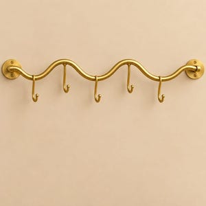May include: A wall-mounted gold-toned metal coat rack featuring a continuous wavy horizontal bar design. The rack includes five downward-curving hooks attached to the bar and two circular mounting plates at each end with visible screw holes.