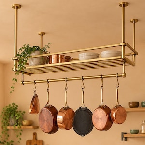 Puede incluir: Un estante para ollas de latón montado en el techo con una repisa rectangular que sostiene una planta, utensilios de cobre y cuencos blancos. Debajo, una barra horizontal con seis ganchos en forma de S sostiene sartenes de cobre y metal.