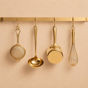 May include: A gold-toned metal wall-mounted rack holding four hanging kitchen utensils: a round mesh strainer, a deep soup ladle, a small saucepan, and a wire whisk. Each utensil features a polished gold finish and is suspended by a matching hook.